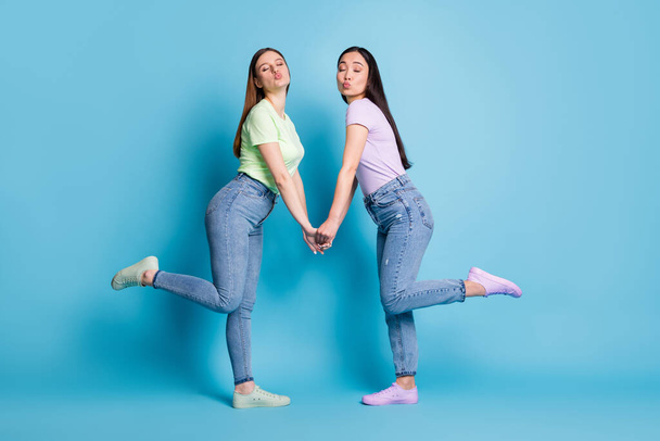 Full size profile photo of two affectionate lesbians couple young students fellowship hold arms send air kisses flirty wear casual t-shirts jeans footwear isolated blue color background - Foto, Bild