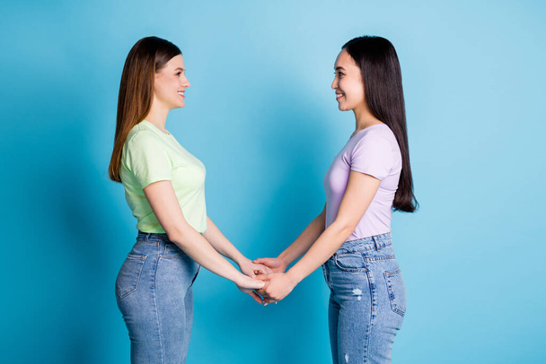Profiel foto van twee aanhankelijke lesbiennes paar jonge studenten beste vrienden vrienden handen kijken ogen gevoelens dragen casual t-shirts jeans schoeisel geïsoleerde blauwe kleur achtergrond - Foto, afbeelding