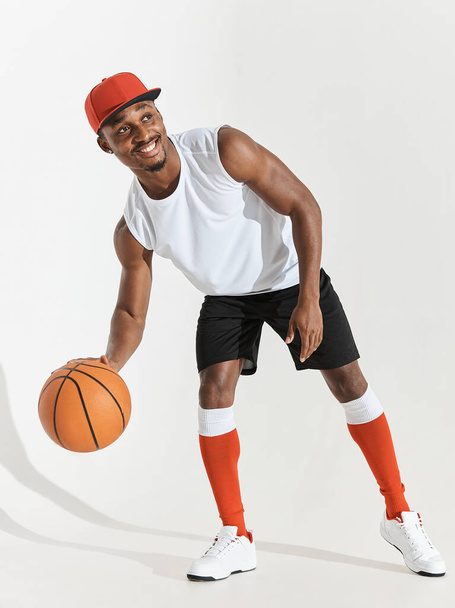 full length photo of a black athletic basketball player in studio on a white background posing with a ball, wearing a white t-shirt, black shorts, red long socks and a cap and white sneakers,he smiles - Fotó, kép