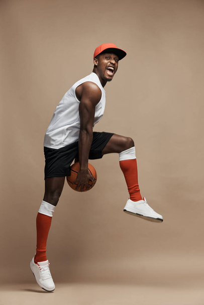 full-length photo of a black  athletic basketball player in the studio on a beige background, jumping high with a ball, wearing white T-shirt, black shorts, red long socks and a cap and white sneakers - Foto, Bild