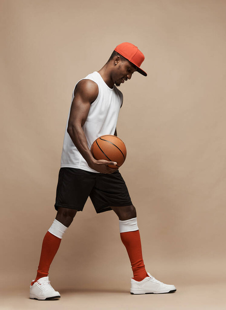 full length photo of a black athletic basketball player in the studio on a beige background wearing white T-shirt,black shorts, red long socks, cap and white sneakers,he holds the ball  and looks down - Foto, Bild