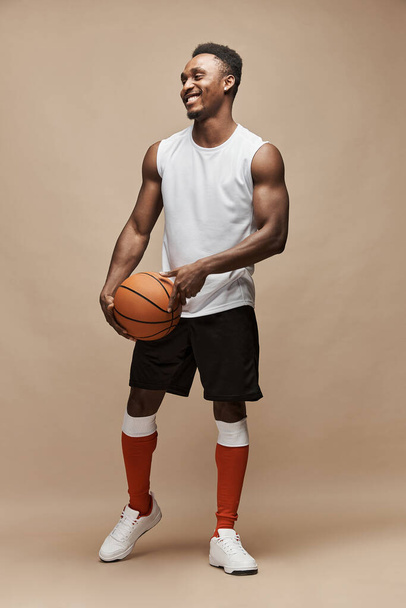 full length photo of a black athletic basketball player in the studio on a beige background wearing a white T-shirt, black shorts, red long socks and white sneakers, he holds the ball and smiles - Zdjęcie, obraz