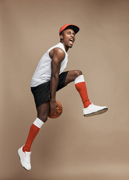full-length photo of a black  athletic basketball player in the studio on a beige background, jumping high with a ball, wearing white T-shirt, black shorts, red long socks and a cap and white sneakers - Fotografie, Obrázek