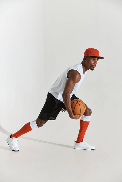 full length photo of a dark-skinned athletic basketball player in studio on a white background posing with a ball, wearing a white t-shirt, black shorts, red long socks and a cap and white sneakers - Fotografie, Obrázek