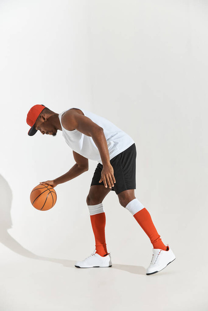 full length photo of a dark-skinned athletic basketball player in studio on a white background posing with a ball, wearing a white t-shirt, black shorts, red long socks and a cap and white sneakers - 写真・画像