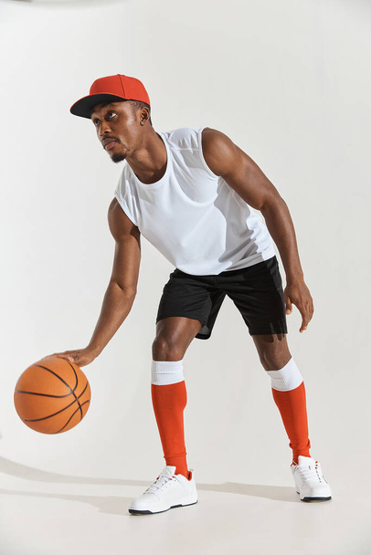 full length photo of a dark-skinned athletic basketball player in studio on a white background posing with a ball, wearing a white t-shirt, black shorts, red long socks and a cap and white sneakers - Zdjęcie, obraz
