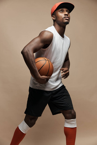 photo of a dark-skinned athletic basketball player in studio on a beige background posing with a ball, wearing a white t-shirt, black shorts, red long socks and a cap, he is looking up  - Photo, Image