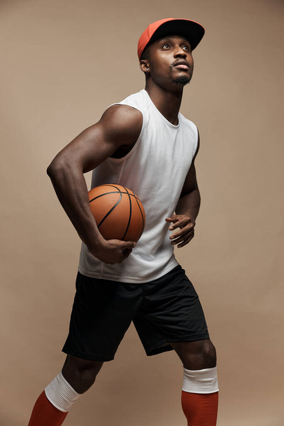 photo of a dark-skinned athletic basketball player in studio on a beige background posing with a ball, wearing a white t-shirt, red cap, black shorts and he is looking up - Zdjęcie, obraz