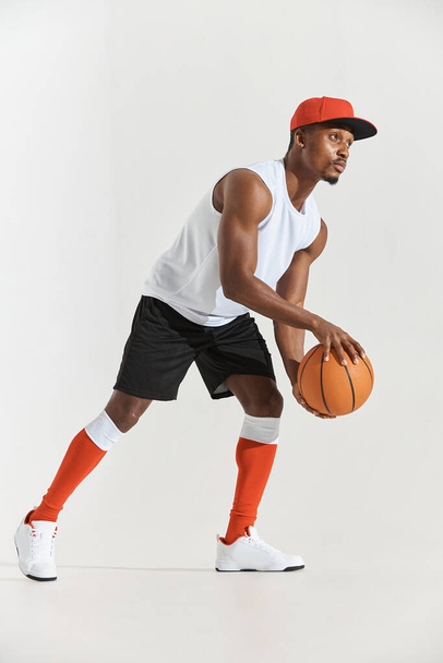full length photo of a dark-skinned athletic basketball player in studio on a white background posing with a ball, wearing a white t-shirt, black shorts, red long socks and a cap and white sneakers - Foto, Imagen