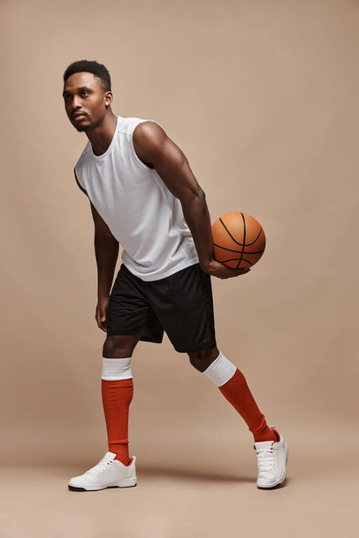 full length photo of a dark-skinned athletic basketball player in studio on a beige background posing with a ball wearing a white T-shirt, black shorts, red long socks and white sneakers - Fotografie, Obrázek