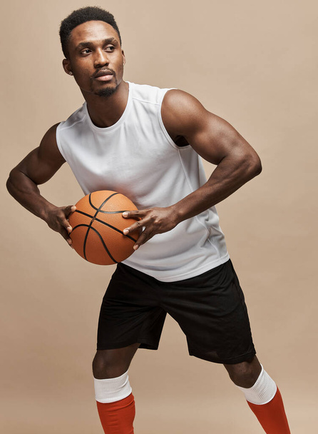 dark-skinned athletic basketball player in studio on a beige background posing with a ball wearing a white T-shirt, black shorts, red long socks  - Foto, Bild