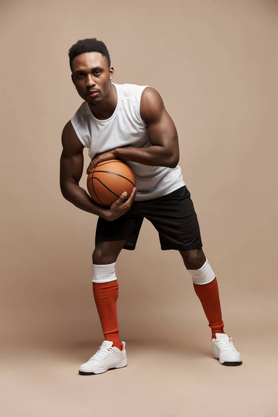 full length photo of a dark-skinned athletic basketball player in studio on a beige background posing with a ball wearing a white T-shirt, black shorts, red long socks and white sneakers - Фото, зображення