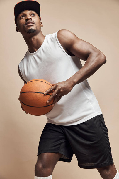 photo of a dark-skinned athletic basketball player in studio on a beige background posing with a ball, wearing a white t-shirt, black shorts, red cap and he is looking up - Foto, afbeelding