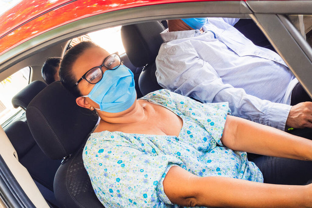 An elderly woman with a medical mask in the passenger seat of the car. Driving to the hospital during the Coronavirus pandemic concept. Road trip, travel and elderly concept - happy elderly couple - Photo, Image