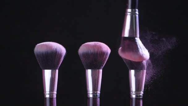 Conceito de beleza. Escovas cosméticas com pó cosmético rosa espalhando no fundo preto em câmera lenta
 - Filmagem, Vídeo