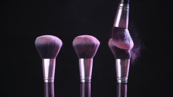 Concepto de belleza. Cepillos cosméticos con polvo cosmético rosa extendiéndose sobre fondo negro en cámara lenta
 - Metraje, vídeo