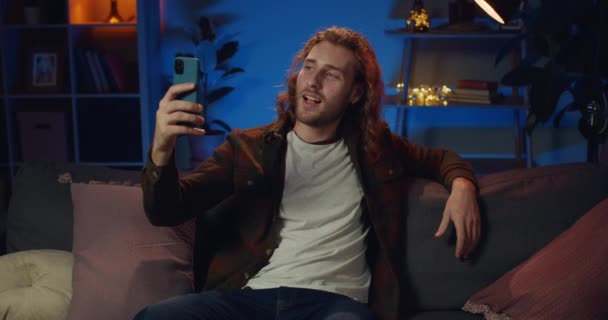 Millennial male blogger smiling and speaking while sitting on couch at home. Cheerful guy gesturing, talking and looking to smartphone camera while doing video chat. Concept of lifestyle. - Metraje, vídeo