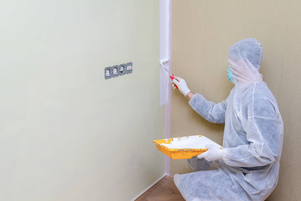 Mies suojaavissa työvaatteissa maalaamassa seiniä valkoisella värillä maalirullalla. Asuntoremontti. Asuntojen uusiminen. - Valokuva, kuva