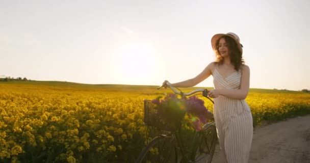 Uma menina está andando ao longo de uma estrada de campo com uma bicicleta. Bicicleta retro bonita com uma cesta de flores. Pôr-do-sol ao fundo. 4K
 - Filmagem, Vídeo
