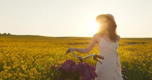 Dziewczyna stoi i opiera się o rower. Patrzy w kamerę i uśmiecha się. Słońce świeci w tle. Podnosi rękę i trzyma kapelusz. 4K - Materiał filmowy, wideo