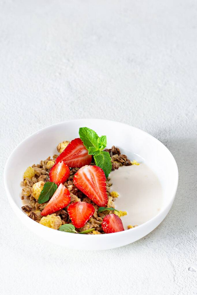 Hausgemachtes knuspriges Müsli mit Nüssen, Trockenfrüchten, frischen Erdbeeren, Minze und Joghurt (fermentierte Backmilch) auf einem weißen Teller. Konzept für gesundes Frühstück. - Foto, Bild