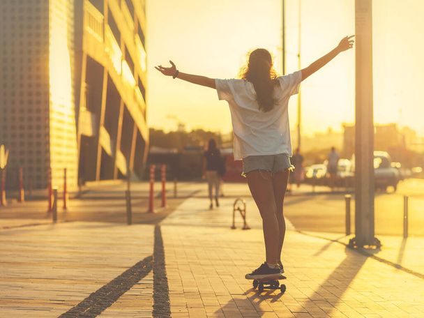 Κορίτσι σε ένα skateboard φωτίζεται από τις ακτίνες του ήλιου που δύει, με τα χέρια ψηλά, την πλάτη της στην κάμερα. Επιλεκτική εστίαση. - Φωτογραφία, εικόνα