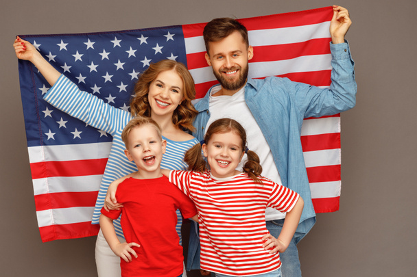 Ευτυχισμένοι γονείς με τη σημαία των ΗΠΑ και χαρούμενα παιδιά αγκαλιάζονται και κοιτάζουν την κάμερα ενώ γιορτάζουν την Ημέρα Ανεξαρτησίας ενάντια στο γκρίζο backgroun - Φωτογραφία, εικόνα
