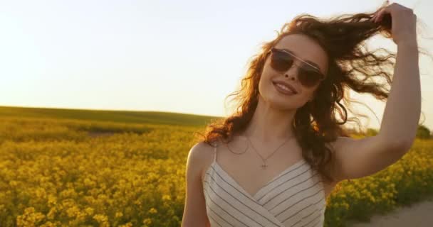 Közelről lőtték le. Egy mosolygó lány sétál a mezőn, és leveszi a kalapját. Megigazítja a haját és leveszi a szemüvegét. 4K - Felvétel, videó