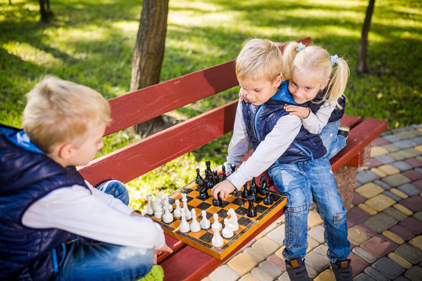 Parktaki bankta satranç oynayan küçük okul çocukları. Yaz tatilleri, entelektüel oyunlar. Çocuklar için satranç kulübü. Mutlu çocukluk ve çocukların arkadaşlığı. Beyin gelişimi için mantık oyunu. - Fotoğraf, Görsel
