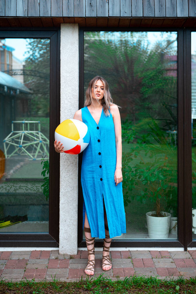 A pretty girl in a summer dress poses against the background of the pool window in the backyard with an inflatable ball. - 写真・画像