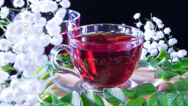 Кружка красного чая с белыми цветами гибискуса и зелеными листьями целебного чая на деревянной подставке. Фото красного травяного индийского целебного чая. Элегантные кружки с расслабляющим и тоником - Фото, изображение