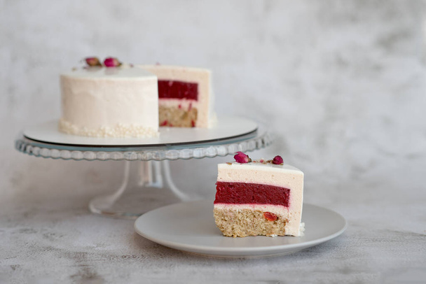 Piece of mousse cake with milk and berry filling  is on a gray simple plate next to the cut cake on  a round transparent glass stand. Cake decorated with dried rose buds.  - Foto, immagini