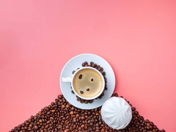 Flat lay. White cup with aromatic coffee on a hill of coffee beans and marshmallows on a pink background. Coffee beans are floating in a cup. Gourmet breakfast. Copy space. - Photo, Image