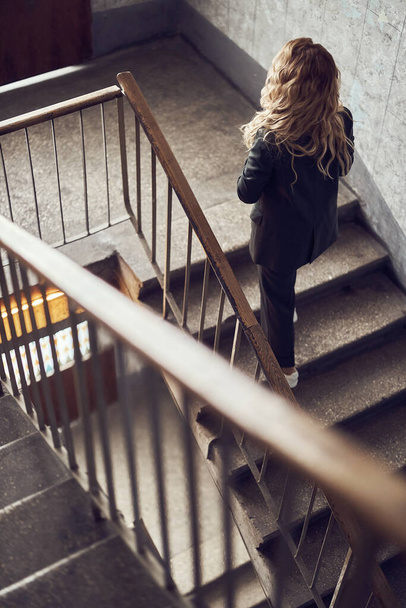 photo of a woman with long blonde curly hair dressed in a black suit climbs the stairs - Fotografie, Obrázek