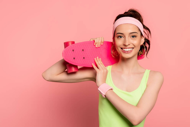 cheerful girl with headband holding penny board on pink  - Фото, зображення