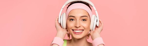 horizontal image of cheerful girl touching headphones while listening music isolated on pink  - Foto, imagen