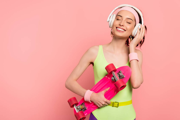 cheerful girl in bodysuit touching headphones while holding penny board isolated on pink  - Fotó, kép