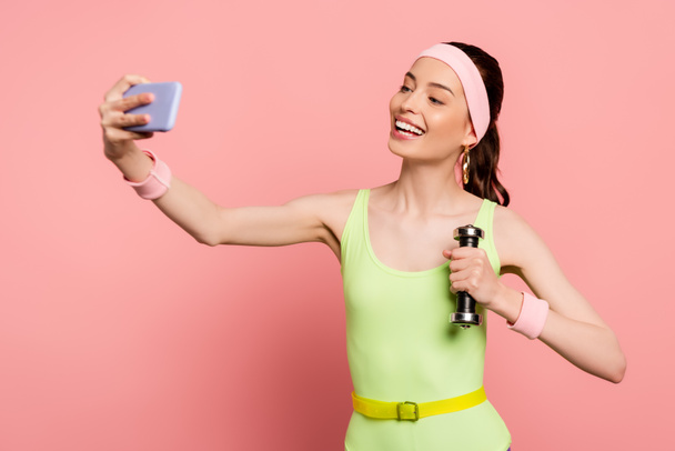 cheerful sportswoman holding dumbbell and taking selfie on pink  - Photo, Image