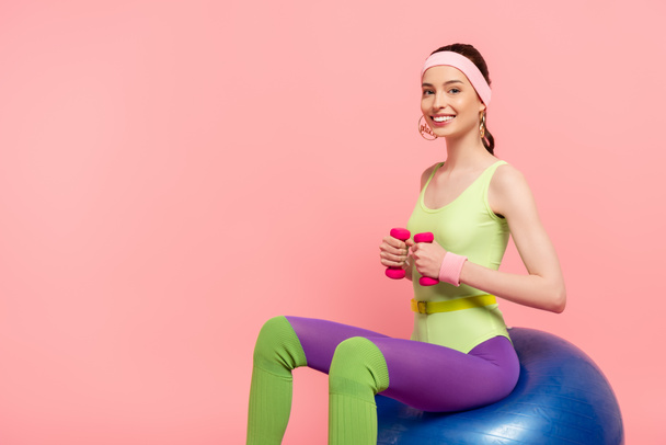 onnellinen nainen istuu kunto pallo ja käyttäessään käsipainot eristetty vaaleanpunainen  - Valokuva, kuva