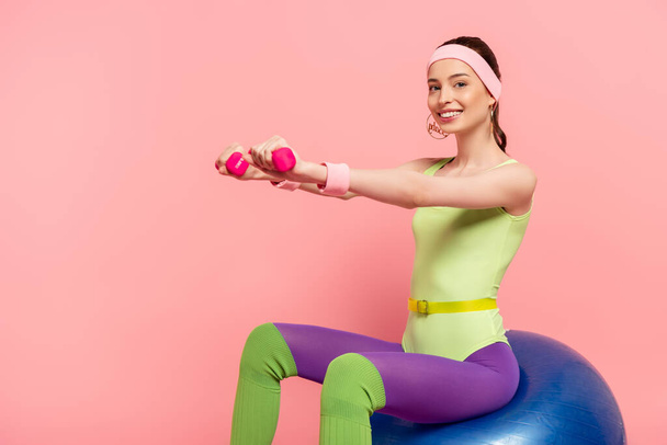 deportista sonriente con las manos extendidas haciendo ejercicio con pesas y sentado en la pelota de fitness en rosa
  - Foto, Imagen