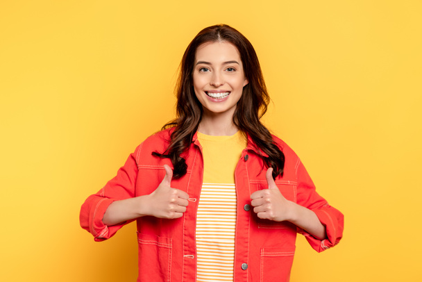 cheerful young woman showing thumbs up and looking at camera on yellow - Foto, afbeelding