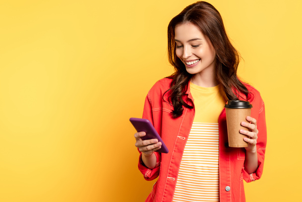 šťastná mladá žena drží kávu jít a pomocí smartphone na žluté - Fotografie, Obrázek