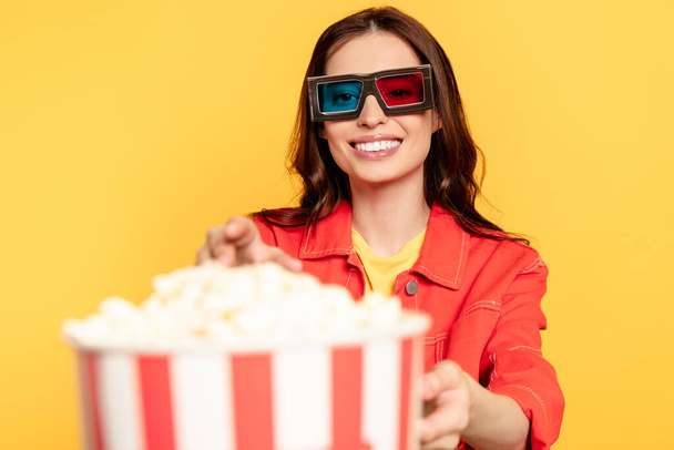 selective focus of happy young woman in 3d glasses reaching popcorn isolated on yellow - Fotoğraf, Görsel