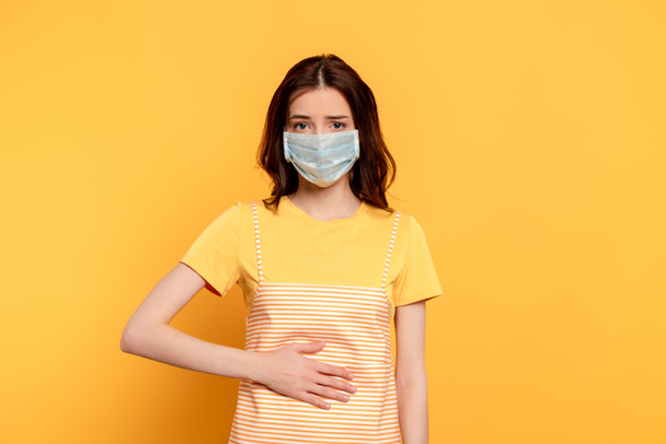 enferma mujer joven en máscara médica que tiene dolor de estómago en amarillo
 - Foto, Imagen