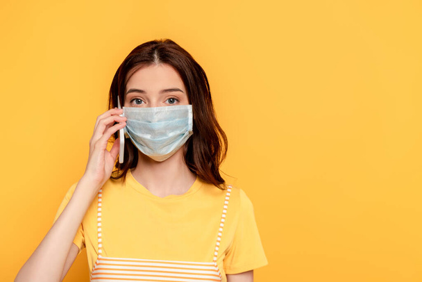 young woman in medical mask talking on smartphone isolated on yellow - Foto, imagen