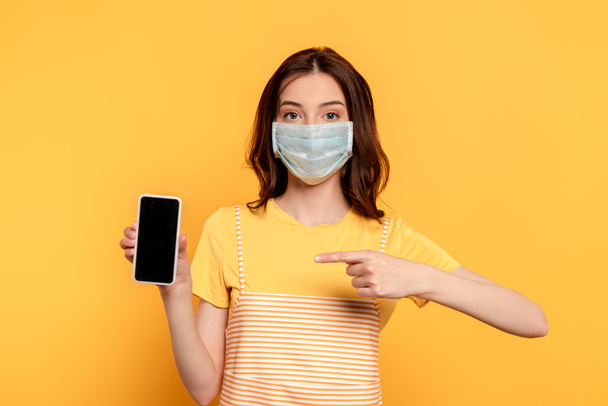 mujer joven en máscara médica apuntando con el dedo al teléfono inteligente con pantalla en blanco en amarillo
 - Foto, Imagen