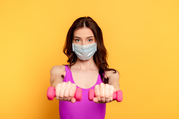 selective focus of sportswoman in medical mask exercising with dumbbells on yellow - Foto, imagen