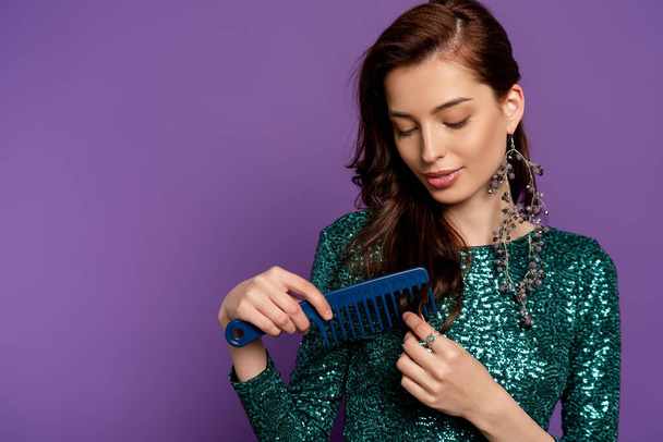 beautiful woman in dress holding comb and brushing hair isolated on purple - Фото, изображение
