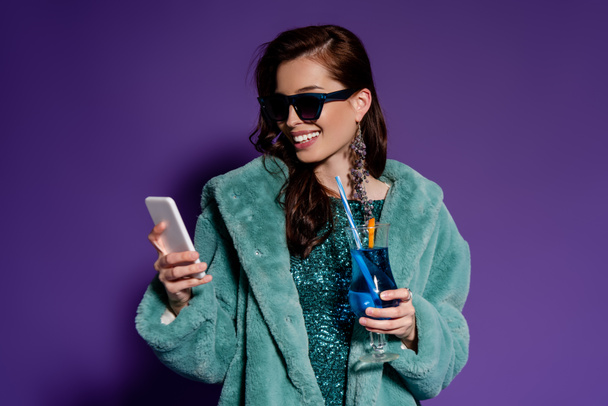gelukkig vrouw in zonnebril holding glas met cocktail en smartphone op paars  - Foto, afbeelding