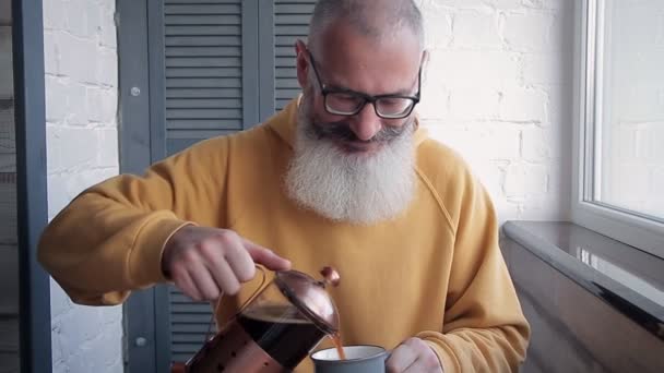 Boldog, érett szakállas férfi túl sok kávét tölt a pohárba, és a kamera elé tárja az italt. Homályos konyhai háttér. - Felvétel, videó